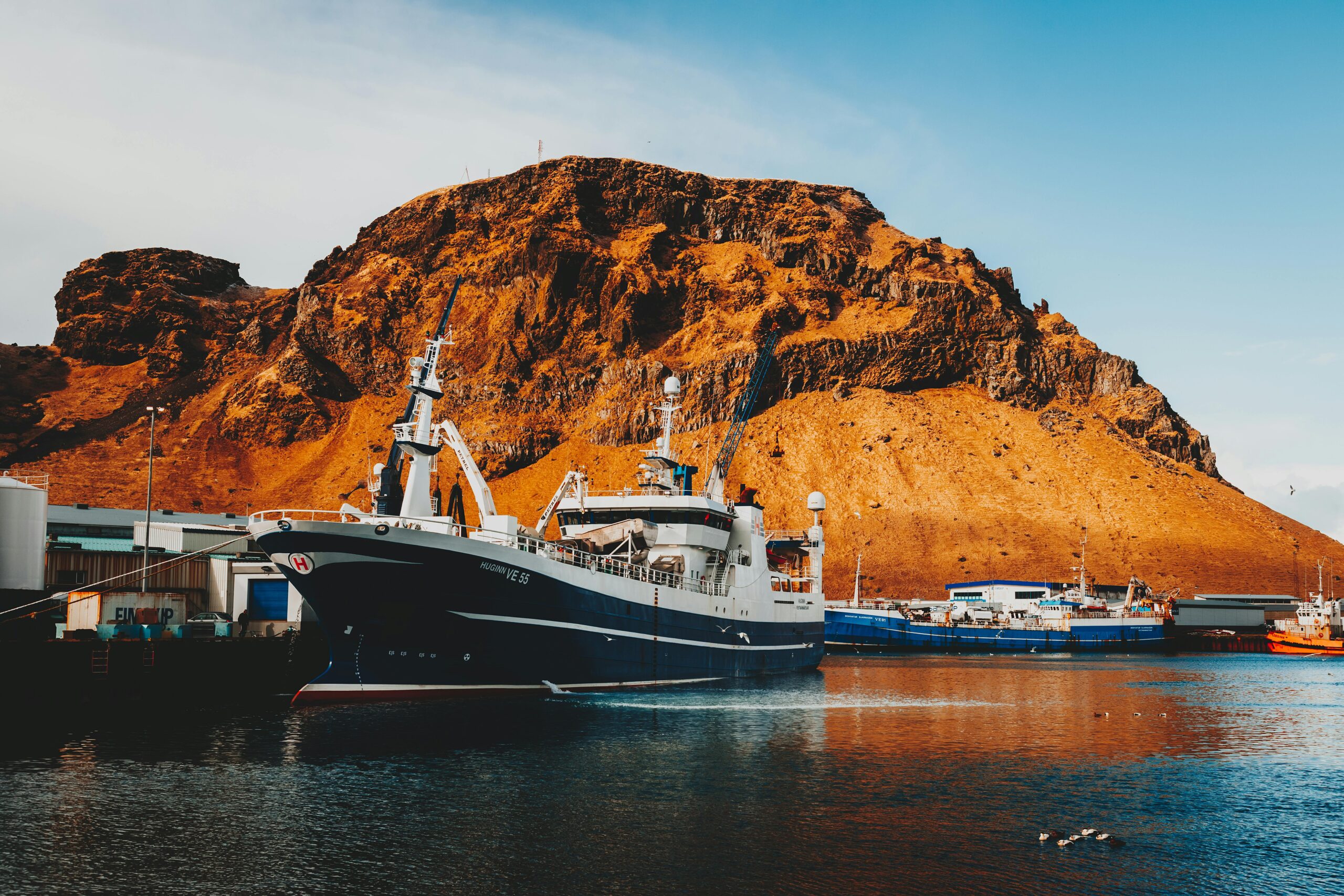 Colorful harbor scene in Vestmannaeyjar featuring ships against a rocky mountain backdrop under a clear blue sky.